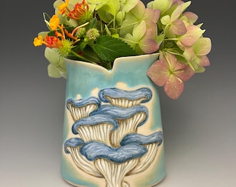 May include: A ceramic vase with a light blue glaze and a design of blue mushrooms. The vase holds a bouquet of flowers with green, orange, and pink blooms. The vase is a unique piece of home decor.