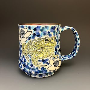 May include: A ceramic mug featuring a hand-painted green frog and black-outlined white flowers set against a background of blue and dark blue dabs of paint. The mug has a matching patterned handle and a smooth, rounded rim with a reddish-brown interior.