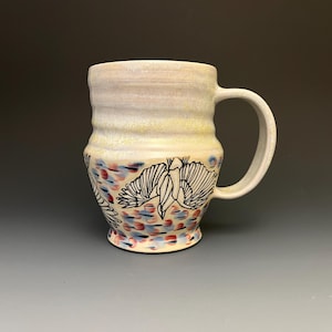 May include: A ceramic mug with a cream-colored glaze and a wide handle. The lower portion features black line art illustrations of birds in flight, set against a background of small, scattered blue and reddish-brown painted brushstroke marks.