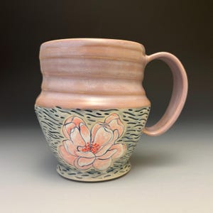 May include: A handmade ceramic mug with a pink glaze and a large floral design. The mug features a light pink handle and a detailed flower illustration in shades of pink and red. The base of the mug has a textured pattern in blue and black.