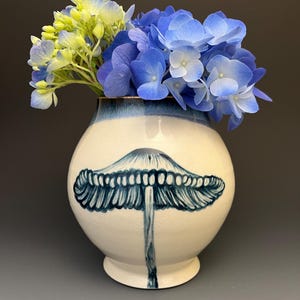 May include: A round ceramic vase with a cream-colored base and a blue-painted mushroom illustration on the front. The rim features a blue glaze, and the vase is filled with a bouquet of vibrant blue and pale yellow hydrangea flowers.