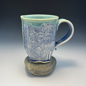May include: A blue and white ceramic mug with a swirling cloud design. The mug has a handle and is sitting on a gray stone.