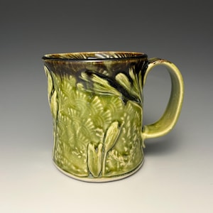 May include: A handmade ceramic mug with a green and brown glaze. The mug features a textured floral design and a curved handle. The interior and handle are a lighter green, while the rim is dark brown.
