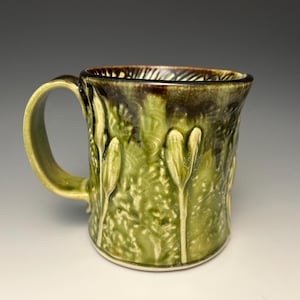 May include: A handmade ceramic mug with a textured, nature-inspired design. The mug features a green and brown glaze, with raised floral or leaf-like patterns. The handle and interior rim are a lighter green, creating a contrast.