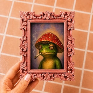 May include: A rectangular, ornate, metallic rose-gold frame containing a print of a green frog wearing a red mushroom cap with white spots. The frog is depicted with a lit cigarette in its mouth, with a thin trail of smoke rising upward.