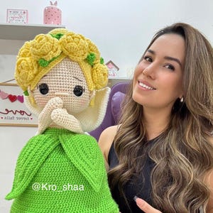 May include: A handmade crochet doll with a yellow floral crown, a green dress, and a white collar. The doll has black eyes and is holding its hands together. The doll's creator's name, "@Kro_shaa", is visible on the green dress.