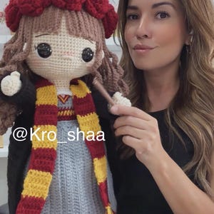 May include: A handmade crochet doll with brown yarn hair, wearing a dark red beret, a black cardigan, a grey dress, and a yellow and dark red striped scarf. The doll has large black eyes and holds a small brown wand. The text @Kro_shaa is visible.