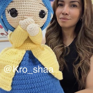 May include: A handmade crochet doll featuring a blue head covering adorned with purple and blue yarn flowers, large black safety eyes, and a beige face. The doll wears a yellow cardigan over a blue dress, with hands held near the face. Text reads @Kro_shaa.