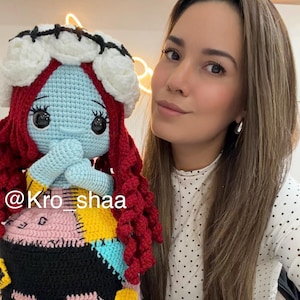 May include: A handmade crochet doll featuring a blue face, red hair, and a patchwork dress. The doll has a white floral headpiece and is holding its hands together. The doll's design is inspired by a character from a popular animated film.