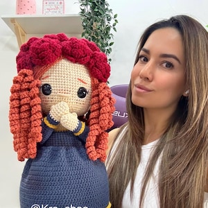 May include: Crocheted doll with a tan face, black eyes, and red and orange curly hair. The doll wears a navy blue dress with a yellow trim. The doll's hands are clasped together. The doll is handmade.