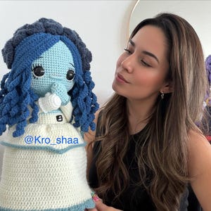 May include: A handmade crochet doll with a blue and white color scheme. The doll has blue hair, a blue face, and is wearing a white dress with blue trim. The doll's design is inspired by a character from a movie. The doll has the text "@Kro_shaa" on the dress.