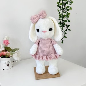 May include: A handmade crochet bunny doll with a pink dress and bow. The bunny has long white ears, black eyes, and a pink nose. It stands on a small wooden base. The doll is a charming, handcrafted item.