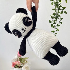 May include: A crocheted panda plush toy with black and white fur. The panda has large black eyes and a black nose. The toy is being held up by a hand.
