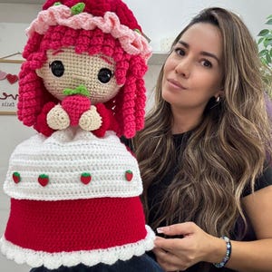 May include: A handmade crochet doll resembling Strawberry Shortcake. The doll features a red dress, white apron with strawberry accents, and a pink bonnet. The doll is holding a small strawberry. The doll's hair is a vibrant pink.