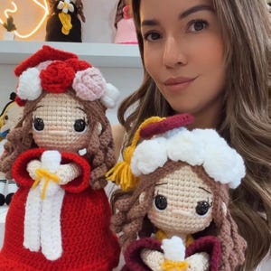 May include: Two handmade crochet dolls featuring brown yarn hair, large black eyes, and graduation attire. One doll wears a red dress with a white floral headpiece, while the other wears a maroon gown with a white cap and a yellow tassel.