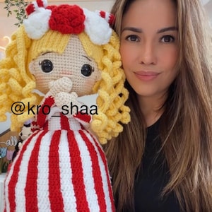 Puede incluir: Muñeca de ganchillo hecha a mano con cabello rizado amarillo, un vestido a rayas rojas y blancas y un lazo a juego. La muñeca tiene ojos negros grandes y junta las manos. El texto "@kro_shaa" es visible.