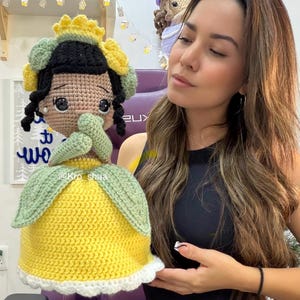 Puede incluir: Muñeca de ganchillo hecha a mano que se asemeja a una princesa, con un vestido amarillo y detalles verdes y blancos. La muñeca tiene cabello negro, una corona amarilla y detalles florales verdes. La cara de la muñeca está detallada con ojos negros y una expresión neutra.
