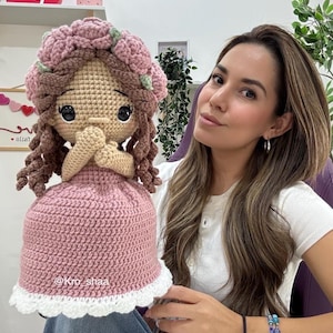 May include: A handmade crochet doll with a pink dress and a floral headpiece. The doll has brown curly hair and is holding its hands together. The dress has a white scalloped trim. The doll is a charming handmade item.