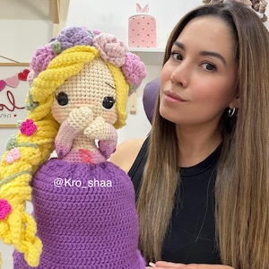 May include: A handmade crochet doll with long yellow hair, a purple dress, and a floral crown. The doll has large black eyes and is holding its hands together. The doll is approximately 12 inches tall. The doll is a handmade item.