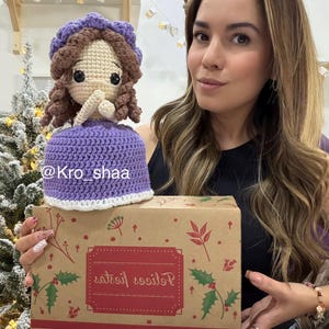 May include: A handmade crochet doll with a purple dress and hat, and brown curly hair. The doll is sitting on a brown box with a red label and Christmas-themed decorations. The text "Kro_shaa" is visible on the doll's dress.