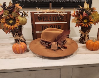 May include: A brown felt fedora hat with a brown leather band and a brown feather. The hat is sitting on a white table with a wooden sign that says "Welcome to our" and two fall-themed arrangements with pumpkins.
