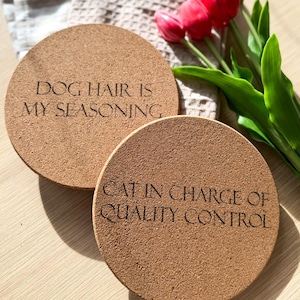 Könnte beinhalten: Zwei runde Untersetzer aus Kork mit eingraviertem Text. Einer lautet "DOG HAIR IS MY SEASONING", der andere "CAT IN CHARGE OF QUALITY CONTROL". Ein gefaltetes beiges Tuch und ein Strauß roter Tulpen mit grünen Blättern befinden sich im Hintergrund.