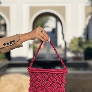 May include: A burgundy macrame handbag with a geometric pattern. The bag has a rectangular shape with a woven design and braided handles. The bag is held up against a blurred outdoor background.