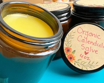 May include: Two amber glass jars with lids, one filled with a yellow salve. The lid of the other jar has a brown paper label with the text "Organic Calendula Salve 4 oz" and a floral design.