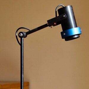 May include: A black desk lamp with a cylindrical head and a blue accent ring. The lamp has a long, adjustable arm and a rectangular base. The lamp is on a red surface.