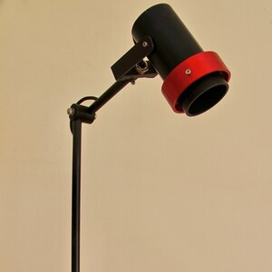 May include: A black desk lamp with a red accent on the shade. The lamp has a flexible arm and a square base. The lamp is plugged into an electrical outlet.