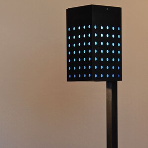 Op de afbeelding: Een zwarte bureaulamp met een vierkante kap met een raster van kleine gaatjes. De kap wordt verlicht door een blauw licht. De lamp heeft een vierkante voet en een hoge, rechthoekige steel.