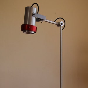 Op de afbeelding: Een zilveren bureaulamp met een rode accent ring op de kap. De lamp staat op een witte gevlekte basis en is geplaatst op een houten oppervlak. Een boek getiteld "An Artist of the Floating World" van Kazuo Ishiguro ligt op de voorgrond.