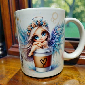 May include: White ceramic coffee mug featuring a cartoon angel with blue wings and a floral crown. The angel is sitting in a coffee cup with a heart drawn on it. The mug has a white handle and is on a wooden surface.