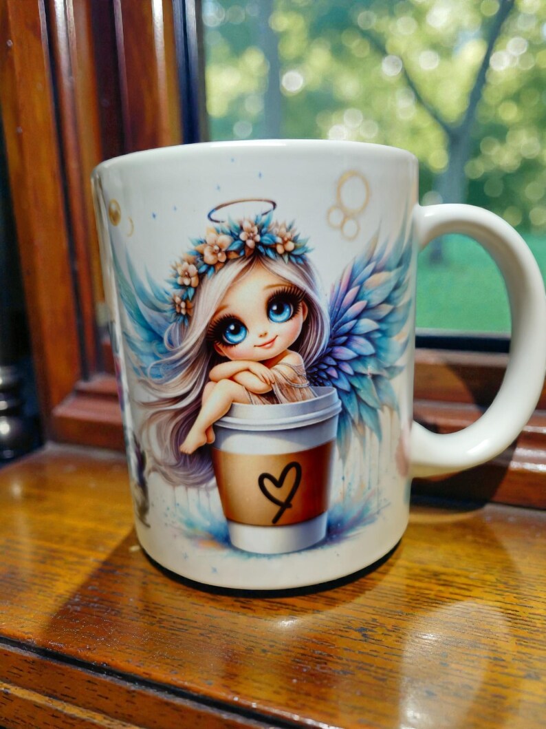May include: White ceramic coffee mug featuring a cartoon angel with blue wings and a floral crown. The angel is sitting in a coffee cup with a heart drawn on it. The mug has a white handle and is on a wooden surface.