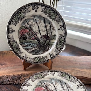 May include: A set of ceramic plates featuring a transferware landscape scene with a central tree, a small house, and a stream. The rim is decorated with a dark brown and green leaf pattern. One plate is displayed on a wooden stand behind a stack of three.