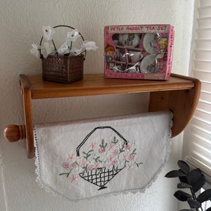 May include: A wooden shelf displays a Peter Rabbit tea set in a pink box, a small woven basket with white flowers, and a white towel with an embroidered floral basket design. The shelf is mounted on a white wall.