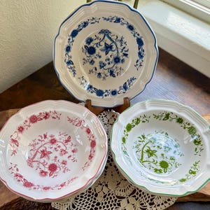 May include: Three decorative ceramic bowls with scalloped edges. One bowl features a blue floral design, another has a red floral pattern, and the third has a green floral pattern. The bowls are white with coloured rims.