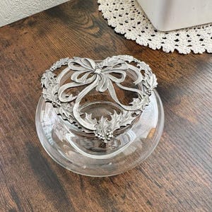 May include: A clear glass bowl with a decorative silver-colored metal wreath and bow design. The wreath features holly leaves and berries. The bowl sits on a dark wooden surface.