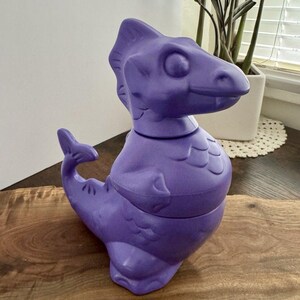 May include: A purple dinosaur-shaped container with a long neck, a fin, and a tail. The container is made of a smooth material and sits on a wooden surface. The dinosaur has a detailed head and body, and the container is likely a toy or decorative item.