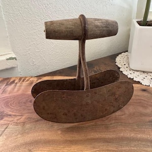 May include: A vintage double-blade mezzaluna herb chopper featuring two curved, rusted metal blades connected by a central metal stem to a horizontal, weathered wooden handle. The tool has a distinct patina showing signs of age and use.