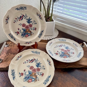 May include: Three off-white ceramic plates with scalloped edges. Each plate features a floral design with pink roses, blue leaves, and a bird. The plates are arranged on a wooden tray, showcasing a vintage aesthetic.