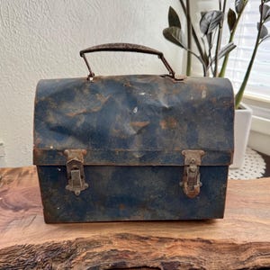 May include: Vintage blue metal lunchbox with a handle and two clasps. The lunchbox shows signs of wear and tear, with rust and dents, giving it a rustic appearance. The lunchbox is sitting on a wooden surface.