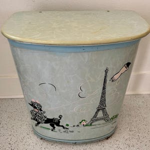 May include: Vintage metal wastebasket with a light blue and white marbled pattern. The wastebasket features illustrations of a poodle, the Eiffel Tower, and a hand reaching for the tower. The lid is light blue with a cream-colored trim.