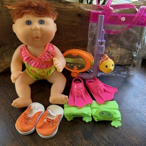 May include: A Cabbage Patch Kids doll wearing a yellow and pink swimsuit, orange shoes, and surrounded by accessories. The accessories include pink flippers, a snorkel, a yellow fish, a mirror, and green turtle-shaped floats, all in a pink tote bag.