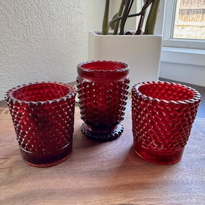 May include: Three ruby red glass candle holders with a hobnail design. The candle holders are different sizes and shapes, with a textured surface. They are arranged on a wooden surface.