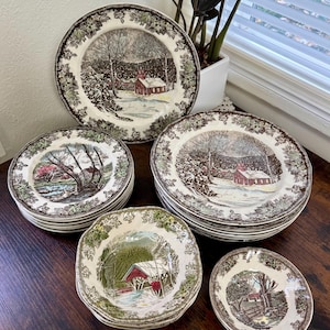 May include: A collection of vintage ceramic plates and bowls with winter landscape scenes. The plates feature a brown and green floral border and a central image of a snowy scene with a red building. Various sizes are stacked on a dark wooden surface.