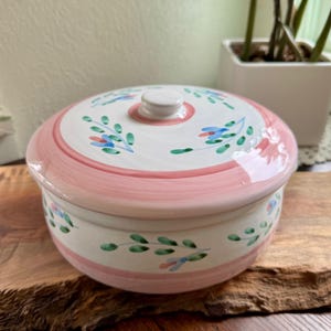 May include: A ceramic lidded serving dish. The white base and lid are decorated with pink stripes and blue and green floral patterns. The dish is on a wooden surface, giving a rustic feel.
