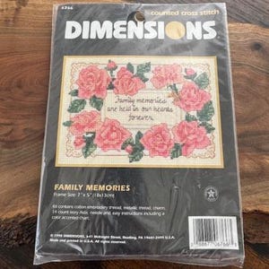 May include: A Dimensions counted cross stitch kit titled "Family Memories." The kit features a cross-stitched design of pink roses and green leaves surrounding the text "Family memories are held in our hearts forever." The frame size is 7" x 5".