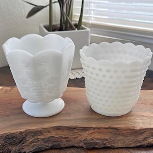 Peut inclure: Deux bols en verre blancs vintage. L'un présente un bord festonné et un motif de raisin et de feuilles. L'autre a un motif hobnail et un bord festonné. Les deux sont sur une surface en bois.