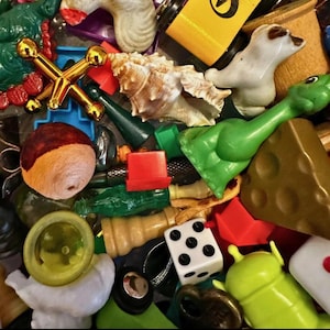May include: A collection of small, colorful toys and trinkets. Includes a gold-colored jacks game, a white ceramic seal figurine, a green plastic dinosaur, a seashell, a white die with black dots, and various other small objects.